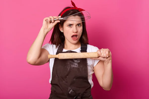 Estudio de aspecto agradable femenina con expresión facial impactada, lleva camiseta blanca, delantal marrón, banda cabezal, sujeción de batidor y rodillo, de pie sobre la pared rosa con espacio vacío . — Foto de Stock