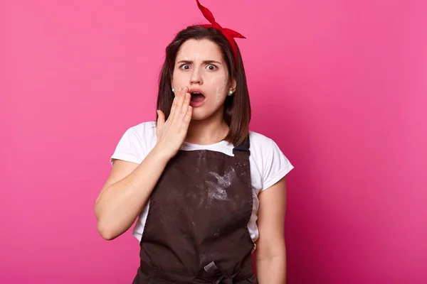 Indoor shot of woman baker wearing brown apron and casual t shirt covers mouth with hand in shock, looks temed, expresando error, olvida añadir ingrediente importante a su pastelería casera . — Foto de Stock