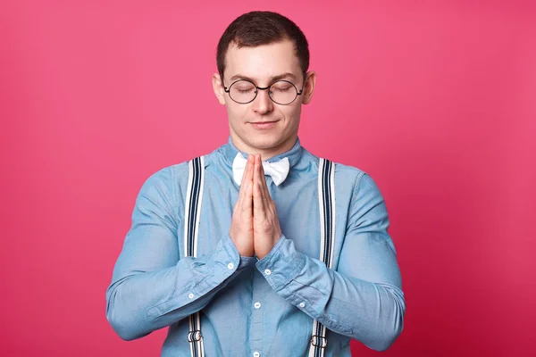 Αθλητικός Ειρηνικός νεαρός άνθρωπος στέκεται με κλειστά μάτια, βάζει παλάμες των χεριών του μαζί, προσεύχεται με χαμόγελο στο πρόσωπό του. Όμορφος ήρεμος τύπος φοράει στρογγυλά γυαλιά, μπλε πουκάμισο, ριγέ τιράντες. — Φωτογραφία Αρχείου