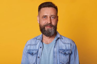Orta yaşlı sakallı gülümseyen sakin adam parlak sarı arka plan üzerinde izole ayakta, doğrudan kameraya bakarak, rahat açık mavi t shirt ve kot ceket giyen, mutlu ve mutlu görünüyor.