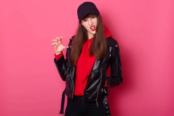 Fashion young teen girl uses bright red lipstick, wearing stylish hoody, leather jacket and black cap. Cheeky tenager poses like tiger. Model poses in studio, looks at camera against pink wall. — Stock Photo, Image