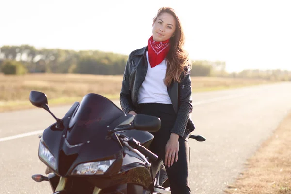 Mooie vrouwelijke bestuurder zit op zwarte snelle Moto Bike, gekleed leren jas en rode bandana, reist rond land door Moto Bike, stopt aan de zijkant van de weg om rust te hebben, kijkt direct naar de camera. — Stockfoto