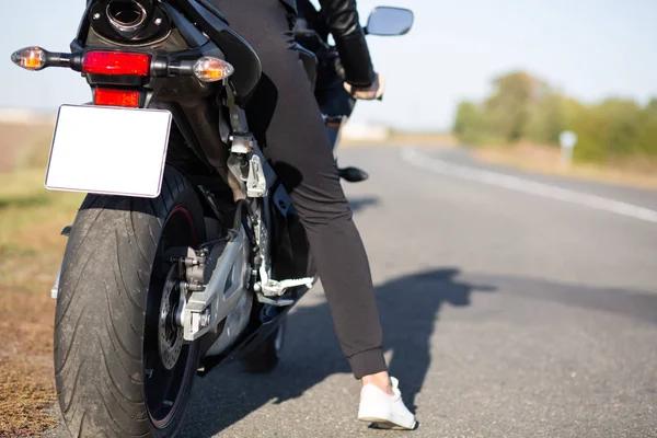 Вид сзади на безликую женщину, сидящую на черном быстром мотоцикле, женщина путешествует одна, отдыхает после долгих часов в пути, дама носит черные брюки отдыхает на обочине дороги. Концепция мототуризма — стоковое фото