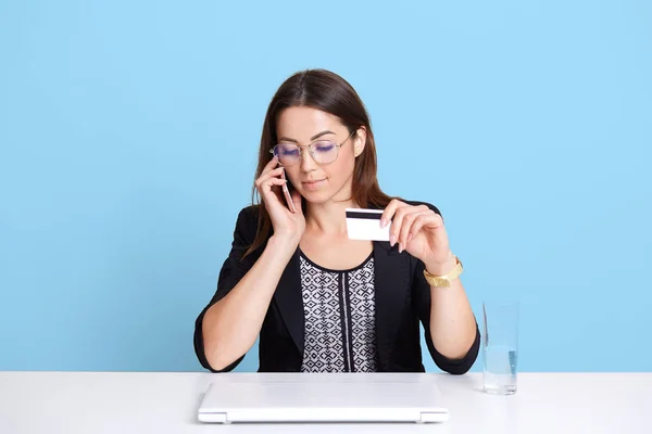 Die junge schöne Geschäftsfrau unterhält sich per Handy, hält die Kreditkarte in der Hand und nutzt elektronische Geräte für den Online-Einkauf, trägt elegantes Hemd und Brille. Einkaufskonzept. — Stockfoto