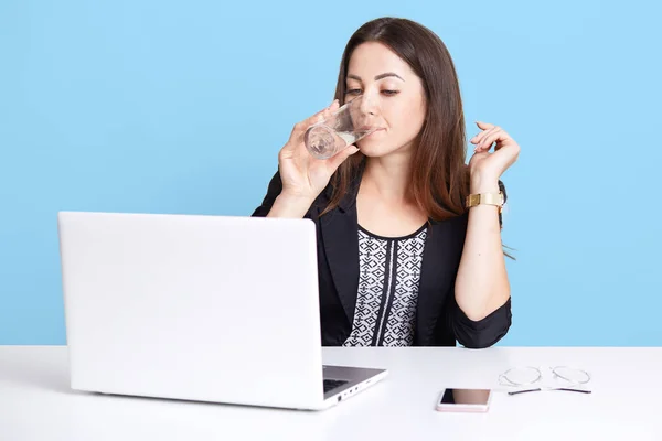 Indoor studio shot of elegant cute pensive young female sitting at office, working hard, drinking water, wearing formal blouse, black jacket and watch, looks serious, isolated over blue background. — Stock Photo, Image