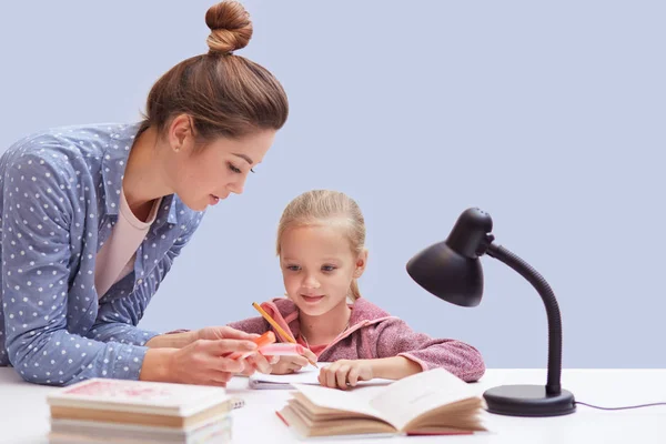 Studio shot of little charming girl sits at table, has difficult homework task, her mother trying to help daughter and explains mathematics rules, uses reading lamp for good vision. Education concept.