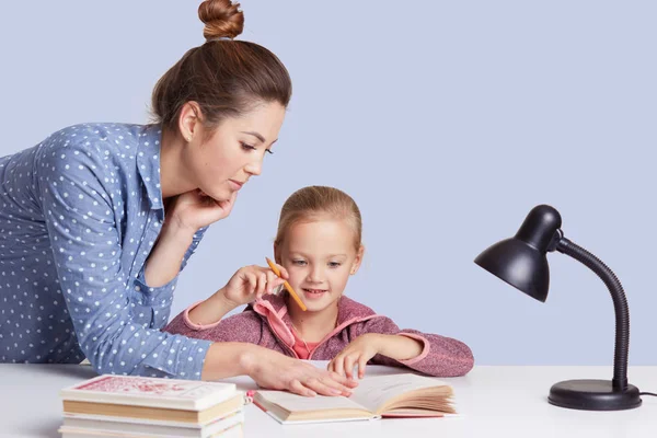 Close up portrait of beautiful Caucasian woman helping her doughter to do school homework, female with bunch on head, wearing casual outfit, little cute girl sitting at table surrounded by books. — Stock Photo, Image
