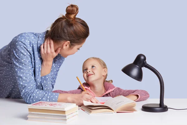 Image of mother and daughter sitting at table surrounded by books looking at each other with love, doing homework together, mummy helps little girl to do sums. Children, school, education concept. — Stock Photo, Image