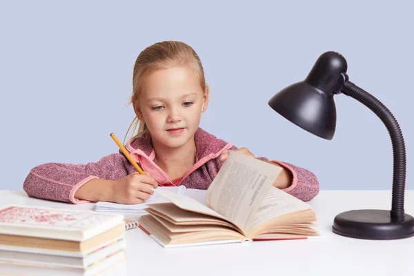 室内拍摄的金发小女孩在白桌看书或写东西，用灯光做功课，已经集中了神情，在演播室里摆姿势，靠着蓝色的墙。教育理念. — 图库照片