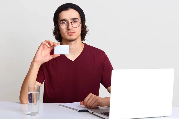 Estúdio tiro de homem sentado na mesa com laptop aberto, estende a mão com cartão em branco para o seu texto de propaganda ou promoção, usa dispositivo eletrônico para pagamento online. Conceito de tecnologia . — Fotografia de Stock