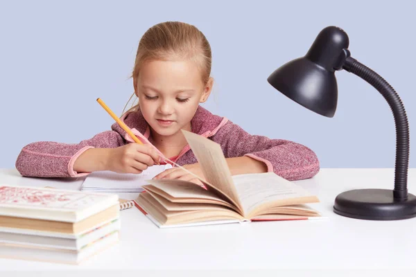 特写画的迷人小女孩坐在白桌前，做家庭作业，试着写作文或做总结，看起来集中，用阅读灯为好视力，隔离在蓝色墙壁上. — 图库照片