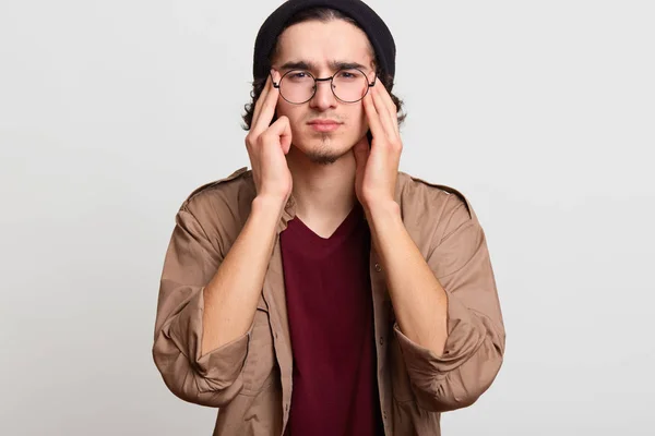 Benadrukte knappe jongere het aanraken van zijn tempels met beide handen, gevoel hoofdpijn, proberen om het te verminderen, ziet er boos. Zwart haired slank model draagt zwarte hoed, rood en beige shirts, brillen. — Stockfoto