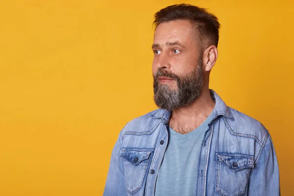 Close up portrait of serious attentive man looking aside, being calm and pensive, wearing casual jeans jacket and blue t shirt, standing isolated over yellow background. Copyspace for advertisement. — Stock Photo, Image