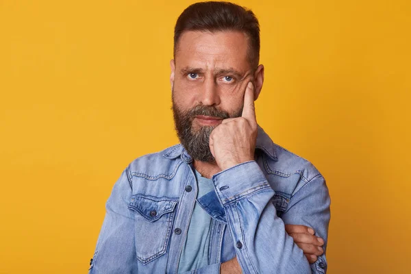 Indoor studio shot of serious magnetic blue eyed man posing isolated over bright yellow background, wearing casual jeans jacket, holding one hand close to face, looks exhausted. Emotions concept. — Stock Photo, Image