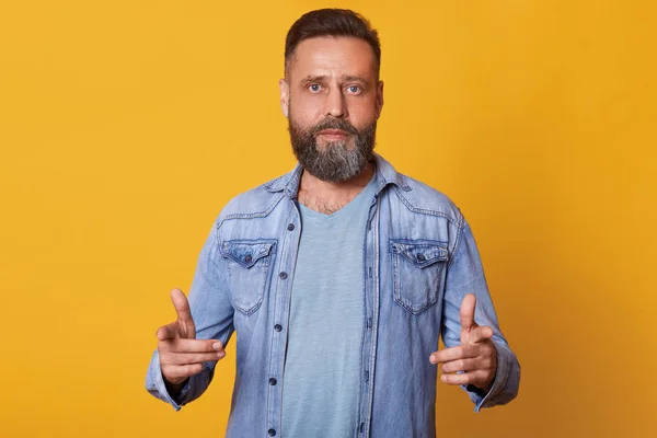 Скромный красивый бородатый мужчина, плетет светло-голубую футболку, джинсовую куртку, показывает направление перед собой двумя указательными пальцами. Уверенная модель среднего возраста, позирующая изолированно на желтом фоне . — стоковое фото