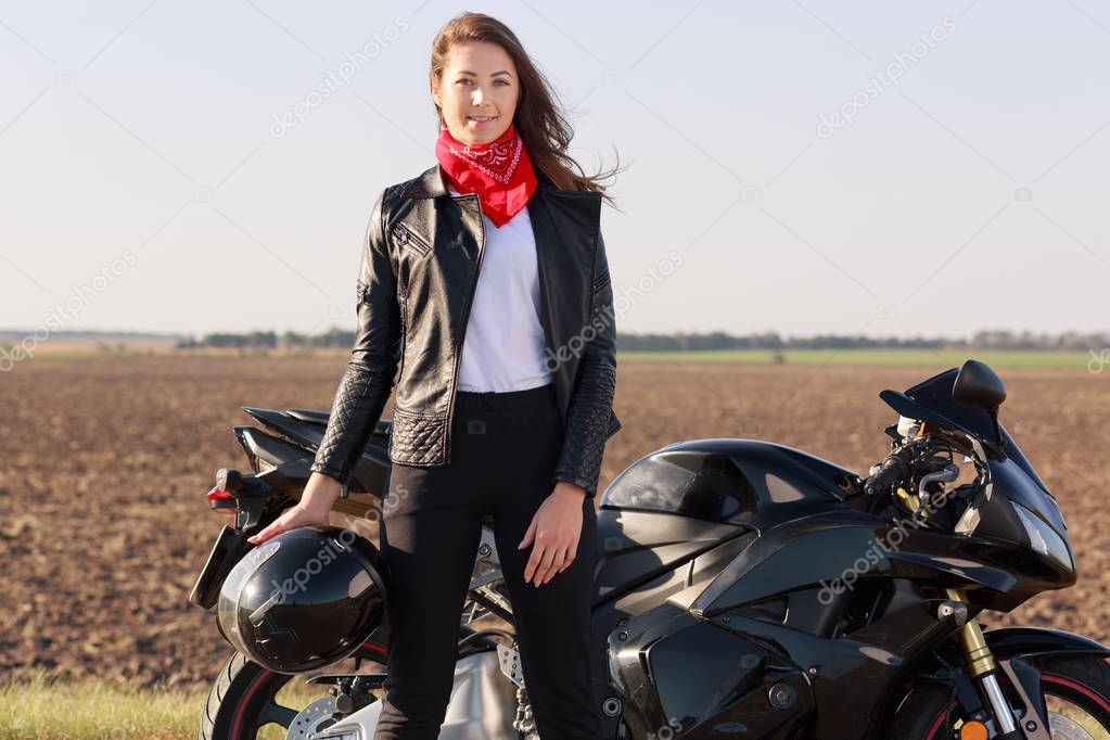 Outdoor shot of attractive woman driver with dark hair stands near black fast motobike, holds helmet, wears leather jacket and red bandana, has confident facial expression. Motorcycle tourism concept.