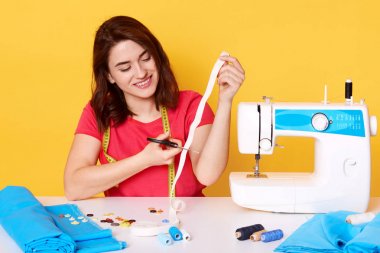 Kanalizasyon mutlu ifade ile oturan kırmızı rahat t shirt giyer, beyaz kurdele kesme, dikiş ekipmanları etrafında poz, onun atölyesinde modern giysiler diker, sarı izole.