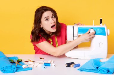 Genç bayan fotoğraf onu dikiş alanında çalışır, dikiş makinesi ile ilgili sorunlar var, aksesuarları oluşturur, giysi tamir, nakış ve paketler trendy giysiler, sarı stüdyo duvarına karşı oturan.