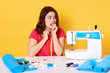 Genç dikiş ağaçlarının yakın çekim tırnaklarını ısırır, dikiş makinesinde sorun yaşıyor, gelişigüzel giyiyor, kenara bakıyor, ne yapacağını bilmiyor, atölyede kanalizasyon çalışıyor. Moda, dikiş konsepti.
