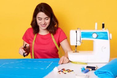 Esmer kız Görüntü kırmızı t shirt giyer, sarı arka plan üzerinde izole oturan, elinde makas tutar, büyüleyici bir gülümseme vardır, şık elbise diker, farklı dikiş ekipmanları ile çevrili oturan.
