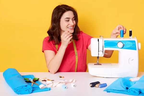 Крытый снимок очаровательной улыбающейся женщины, надевающей нитку на швейную машинку, находящейся в хорошем настроении, ей нравится ее работа, женщине, работающей в мастерской, создающей стильную одежду, изолированной на желтом фоне . — стоковое фото