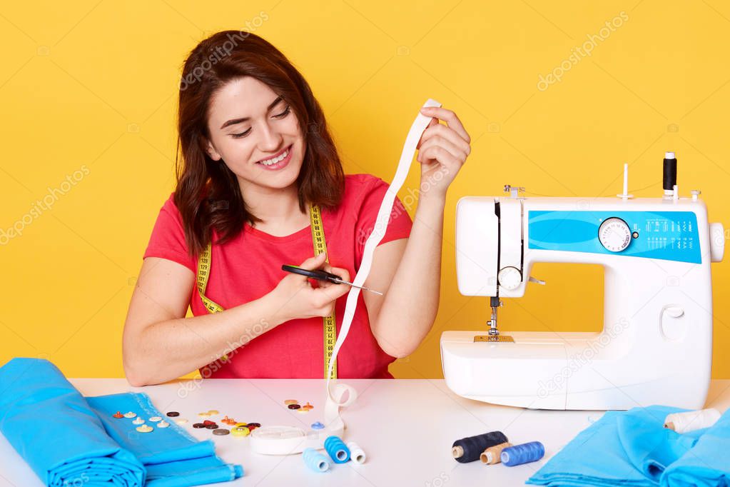 Sewer wears red casual t shirt with happy expression sitting with happy expression, cutting white ribbon, posing around sewing equipment, sews modern clothes in her workshop, isolated on yellow.