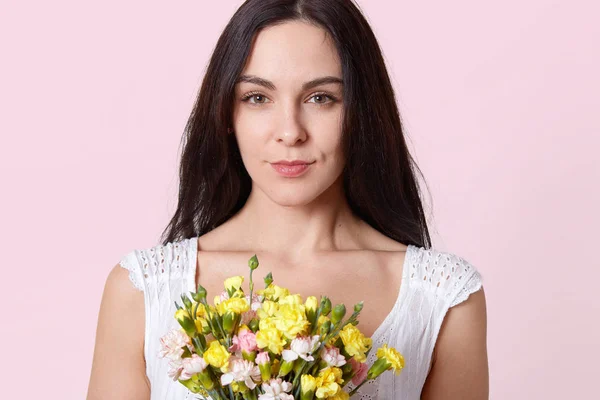 Retrato de moça morena gentil, vestida de vestido branco, recebe presente no aniversário, sendo fotografada com expressão facial séria, segura buquê de flores bonitas, olhando para a câmera . — Fotografia de Stock