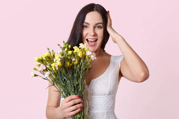 Retrato de meio comprimento de mulher morena bonita segurando um monte de flores, rindo enquanto olha diretamente para a câmera, mantém a mão na cabeça, vestido de vestido branco, isolado sobre fundo de estúdio róseo . — Fotografia de Stock