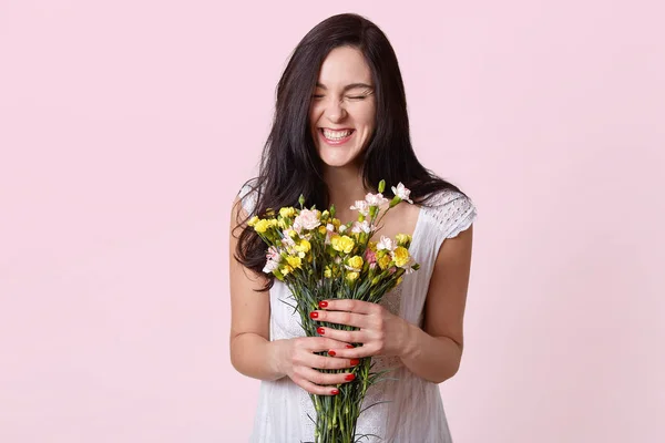 Chica en vestido blanco disfruta del aroma de las flores y se ríe sinceramente, disfrutando de un gran día de primavera sobre un fondo rosa, posando con los ojos cerrados, estando en alto espíritu, siendo feliz en el día de su boda . —  Fotos de Stock