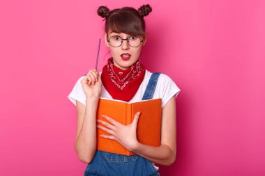 Yaratıcı sevimli kız öğrenci stüdyoda pembe arka plan üzerinde izole duruyor, ağzını ve gözlerini yaygın olarak açar, bir elinde kalem ve diğer bir turuncu defter tutarak, yeni bir fikir sahip. Eğitim kavramı.