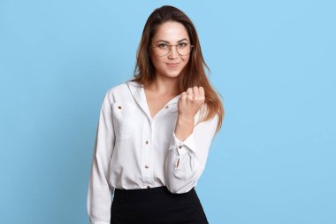 Korkunç zarif patronluk yöneticisi sıkıca ayakta, sıkıştırılmış el tutarak, yumruk gösteren, doğrudan kameraya bakarak. Uzun saçlı kendinden emin modeli beyaz resmi bluz ve siyah etek poz, gözlük.