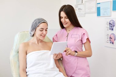 Pembe laboratuvar önlüğü ve beyaz havlu içinde müşterisi, güzellik dolabında beyaz duvarın üzerinde poz veren, birbirleriyle konuşan, mutlu görünen uzun saçlı gülümseyen güzellik uzmanı. Kozmetoloji kavramı.