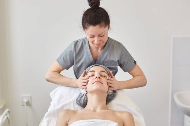 Güzel genç kadın güzellik spa yüz masajı ile rahatlatıcı rahat couh yatarken, gri tıbbi kıyafet içinde messeur müvekkiline masaj. Güzellik, cilt bakımı ve kozmetoloji konsepti.