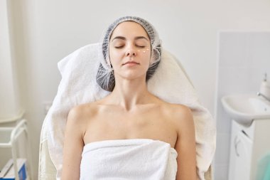 Close up portrait of well kept beautiful woman laying on coach with closed eyes, having peaceful facial expression, being naked, rolled with white towel and medical cap, cream points under eyes. clipart