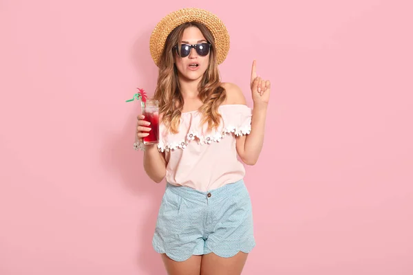 Привлекательная молодая женщина в летнем наряде изолирована на розовом фоне. Счастливая девушка в солнечных очках позирует в соломенной шляпе и коктейле, проводит жаркие летние дни на пляже, указывает пальцем вверх, имеет новую идею — стоковое фото