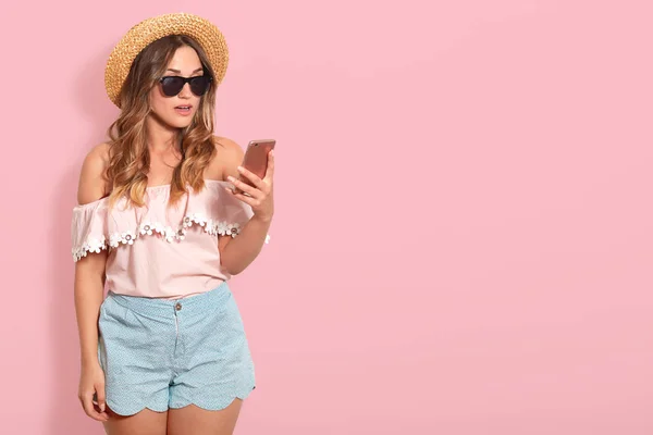 Portrait of astonished pretty woman wearing blouse with bared shoulders, short, straw hat and sunglasses, holds smart phone with surprised facial expression, checking social network, has shocking news — Stock Photo, Image