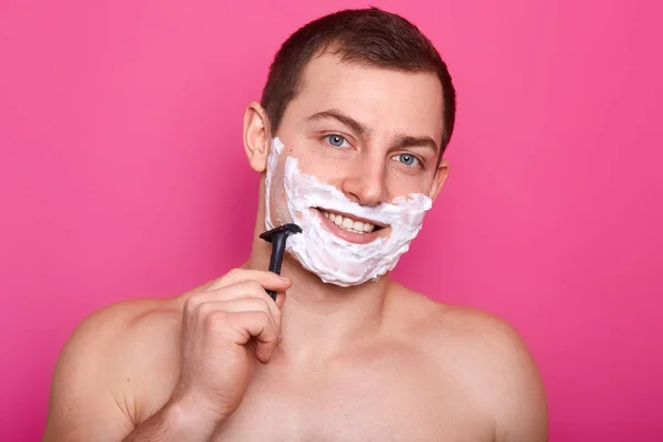 Foto av bar överkropp ung man rakning hans ansikte och titta på kameran medan du står isolerad på rosa bakgrund, poserar naken med leende, att vara i badrum på morgonen, tar hand om sin hud. — Stockfoto