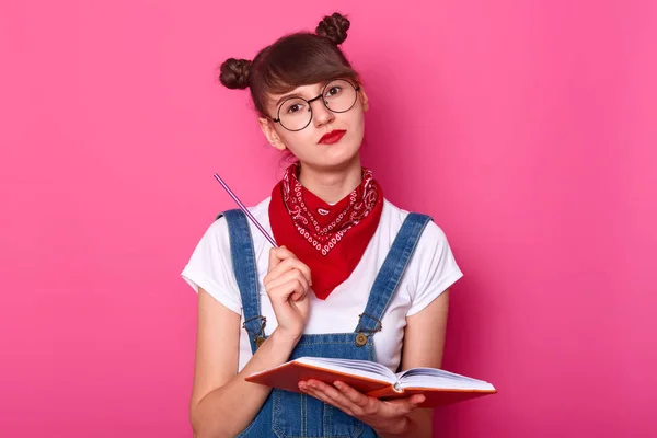 Εσωτερικό στούντιο shot του σκεπτικού στοχαστικό νεαρό μαθητή με κόκκινο κραγιόν στο πρόσωπό της, κρατώντας σημειωματάριο και στυλό στα χέρια της, κάνοντας μερικές σημειώσεις, φορώντας φόρμες, t shirt, κόκκινη μπαντάνα και γυαλιά. — Φωτογραφία Αρχείου