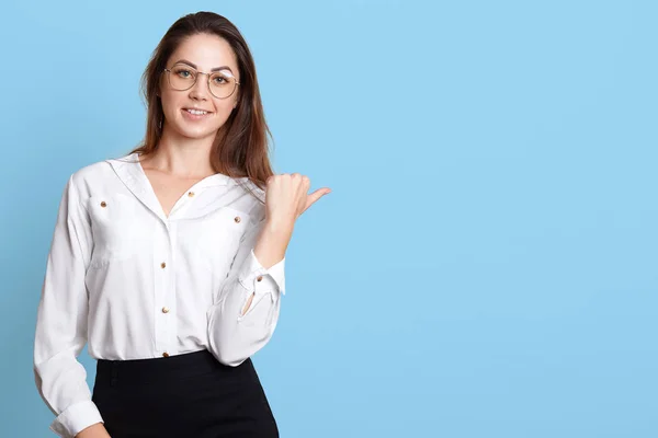 La joven exitosa y segura mira directamente a la cámara, mostrando la dirección con su pulgar, con sonrisa en la cara, usando blusa blanca, falda negra y gafas elegantes. Copiar espacio para publicidad . — Foto de Stock