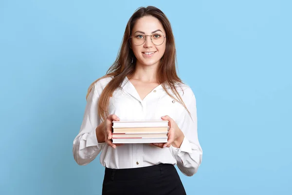 Sonriente secretaria energética delgada sosteniendo pila de libros en ambas manos, habiendo complacido la expresión facial, mirando directamente a la cámara, vistiendo falda negra, blusa blanca inteligente y gafas redondas . —  Fotos de Stock