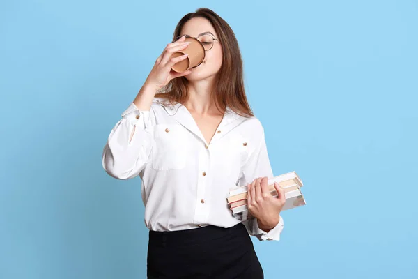 Studio intérieur plan d'un travailleur de bureau mince et travailleur ayant une pause café, boire une boisson chaude à partir de papier tasse, tenant un tas de livres dans une main, posant isolé sur fond bleu clair . — Photo