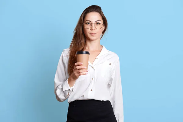 Retrato de una mujer de negocios seria, con camisa blanca, falda negra y gafas, tiene el pelo largo y oscuro, sosteniendo una taza de papel de café para llevar, mira a la cámara, aislado en el fondo azul del estudio . — Foto de Stock
