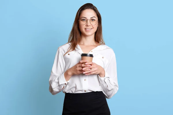 Estúdio tiro de mulher europeia relaxada e sorridente com cabelos longos escuros e óculos, vestindo blusa e saia, segurando takeaway café, desfrutando de grande dia de trabalho isolado sobre fundo azul . — Fotografia de Stock