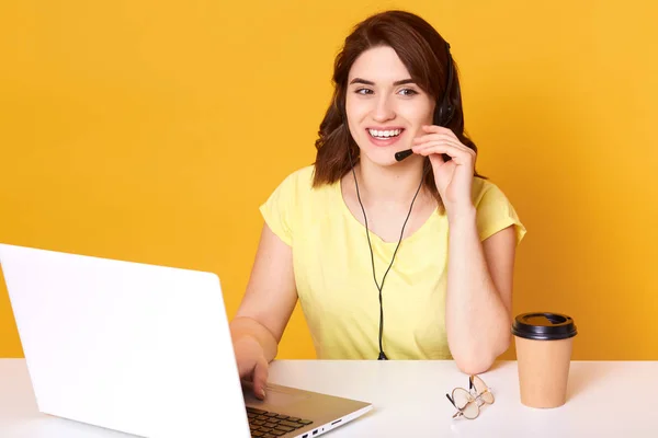 Setengah panjang foto wanita operator duduk di meja kantor, melihat ke samping, menyentuh headset, terisolasi atas latar belakang kuning, wanita berambut cokelat memakai santai, tampak ceria. Konsep call center . — Stok Foto