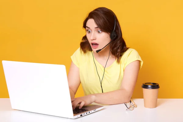 Short haired shocked attractive operator posing with wide opened mouth and eyes, looking at laptop screen, typing some information, having headset, working hard at call center. Work concept. — Stock Photo, Image