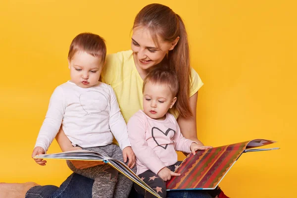 Studio shot of happy family: mother and little twins girls sitting on floor, reading books and viewing bright interesting pictures, spending time with mommy, selebrating Mother's Day. Children concept — Stock Photo, Image