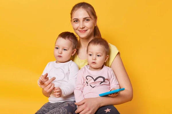 Charming affectionate mummy sitting with her cute little daughters on floor in yellow studio, females dressed casually, looks happy, children spending time with mom, sisters on mommy's knees. — Stock Photo, Image