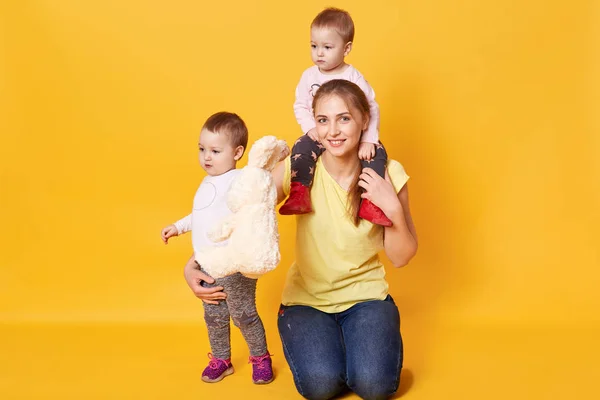 Foto de família feliz. Mamãe com seus gêmeos meninas posando no estúdio, mamãe segura uma criança nos ombros, vestida roupas casuais, sendo fotografada isolada sobre fundo amarelo. Conceito de maternidade . — Fotografia de Stock