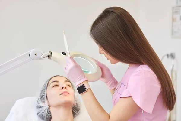 Long haired beautician standing near her client, touching magnifying glass, watching through it, exploring skin condition. Model in medical cap looks at beautician. People and procedures concept. — Stock Photo, Image