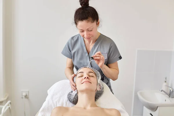 Positive schoonheidsspecialiste werkt bij Beauty Parlor. het hebben van de klant, het toepassen van hydraterende Gezichtsolie, voorbereiding gezicht voor verdere procedures. Ontspannen Glimlachende vrouw die rust in de spa salon. Huid-en verzorgings concept. — Stockfoto